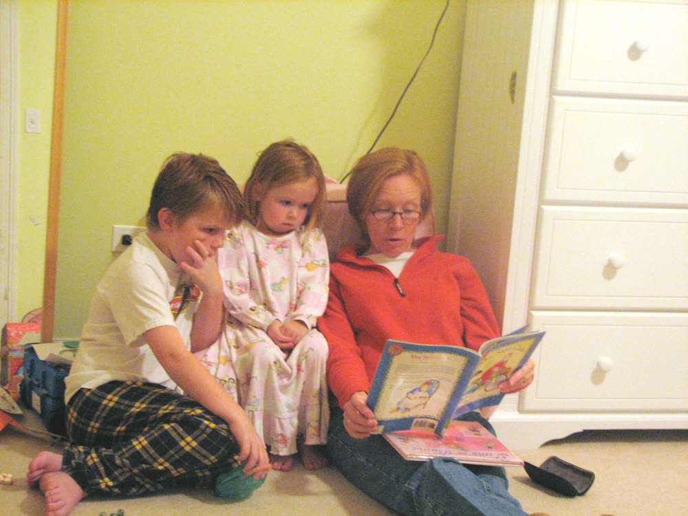 A woman reading a picture book aloud to two children sitting
                  beside her on a bedroom floor